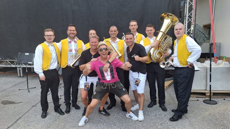 A group of men in yellow vests, some holding musical instruments, pose for a photo on a stage. A man in a pink shirt and sunglasses stands in the middle.