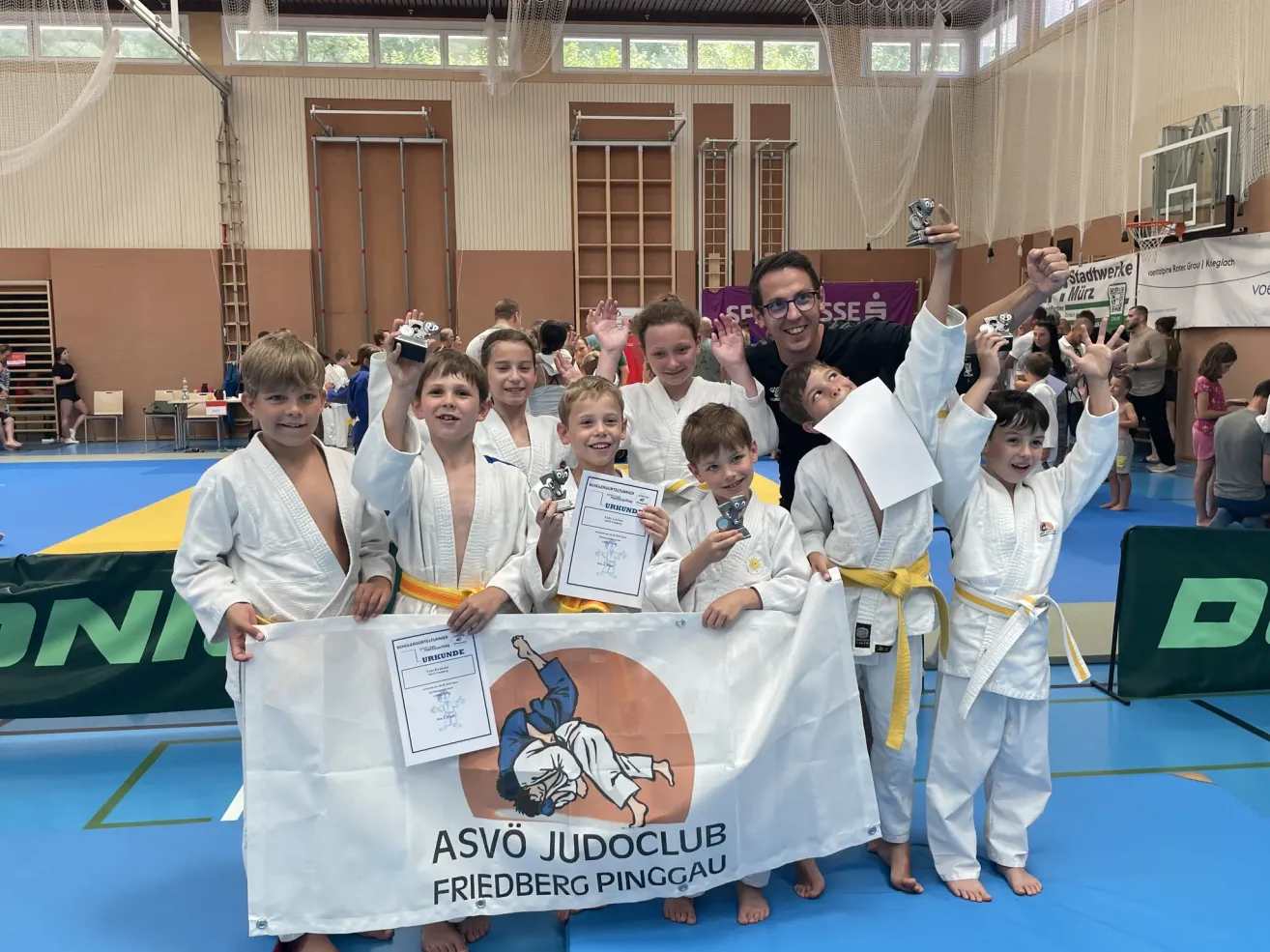 Eine Gruppe junger Judokas in weißen Uniformen mit gelben Gürteln posiert für ein Foto mit Pokalen und Urkunden. Ein Mann mit Brille steht lächelnd hinter ihnen. Ein weißes Banner mit blauem Text und einem Bild eines Judokas wird von den Kindern gehalten.