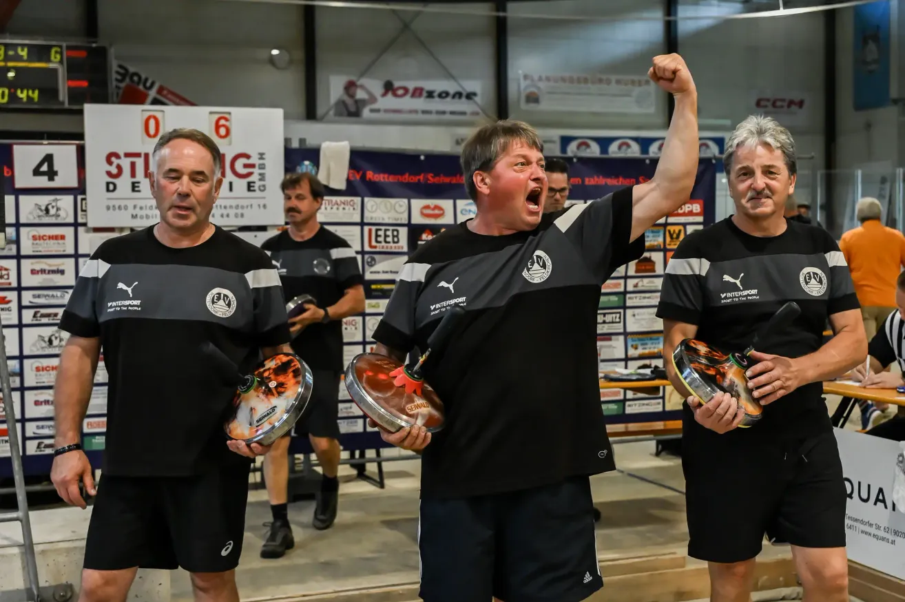 Three men celebrate with trophies in a gym. The central figure raises his fist, while others look on. The room is adorned with banners and promotional materials.