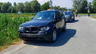 Ein schwarzer BMW SUV mit beschädigter Front ist am Straßenrand geparkt, während zwei Personen in Warnwesten in der Nähe stehen.