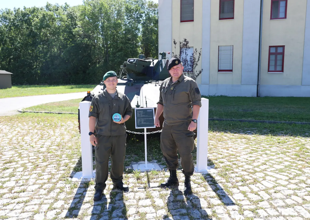 Two men in military uniforms stand in front of an armored vehicle in front of a building.