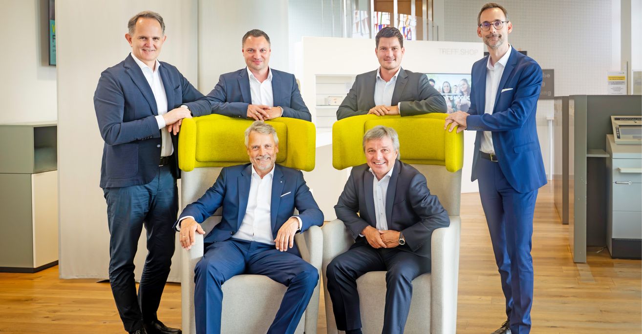 Fünf Männer in Anzügen und Krawatten, die in einem modernen Büro sitzen und stehen und für ein Foto posieren. Zwei Männer sitzen in leuchtend gelben Stühlen, während drei Männer hinter ihnen stehen und lächeln.