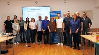 Gruppe von Menschen steht vor einer Präsentationsleinwand und lächelt für ein Foto. Hinter ihnen befinden sich zwei Banner.