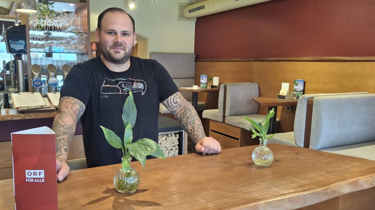 Ein Mann mit Tattoos sitzt an einem Tisch mit Pflanzen in Vasen. Er trägt ein schwarzes Shirt mit einem Seattle Seahawks-Design.