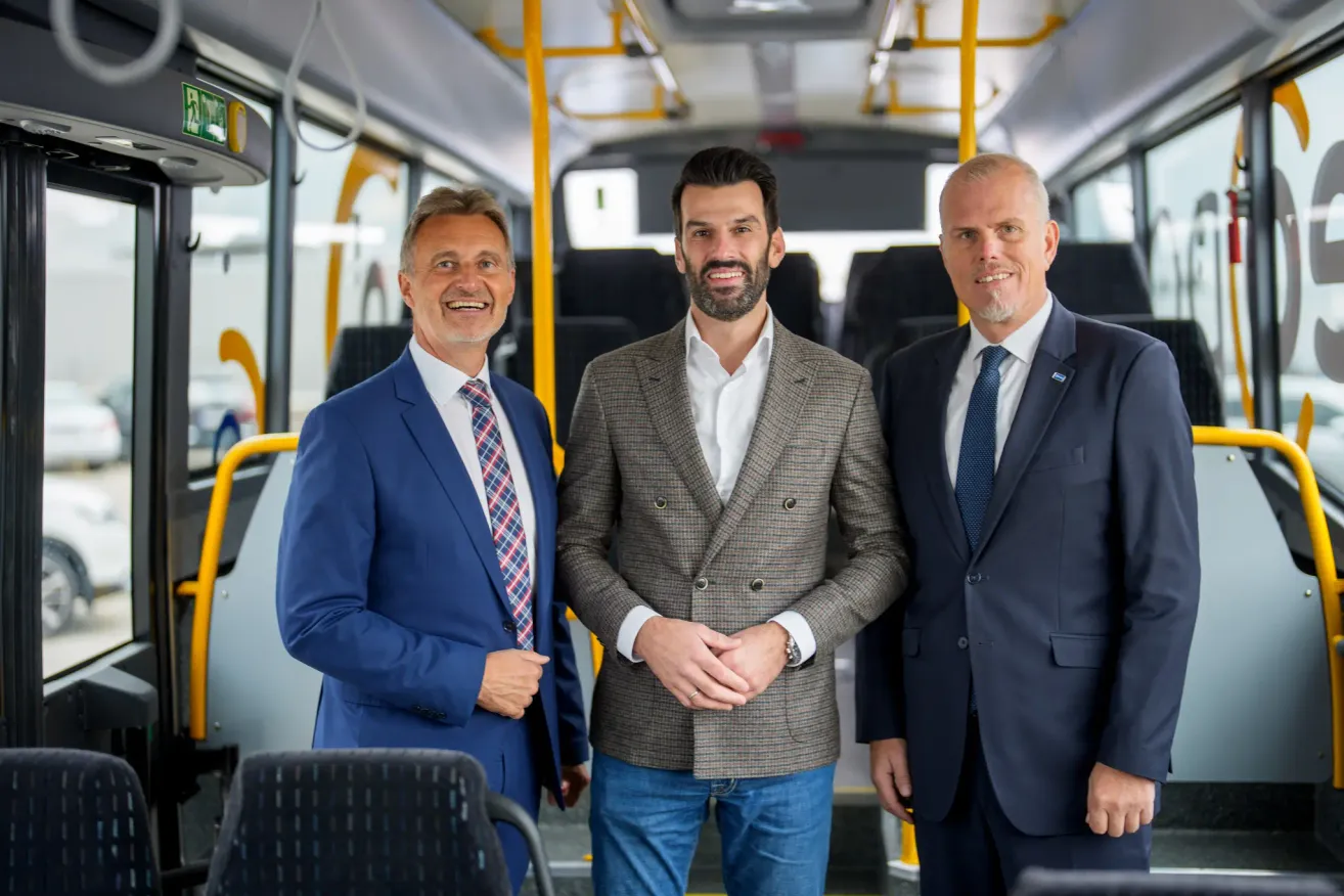 Three men in suits stand inside a bus, smiling for a photo. The man in the middle has a beard and a watch.