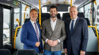 Drei Männer in Anzügen stehen in einem Bus und lächeln für ein Foto. Der Mann in der Mitte hat einen Bart und eine Uhr.