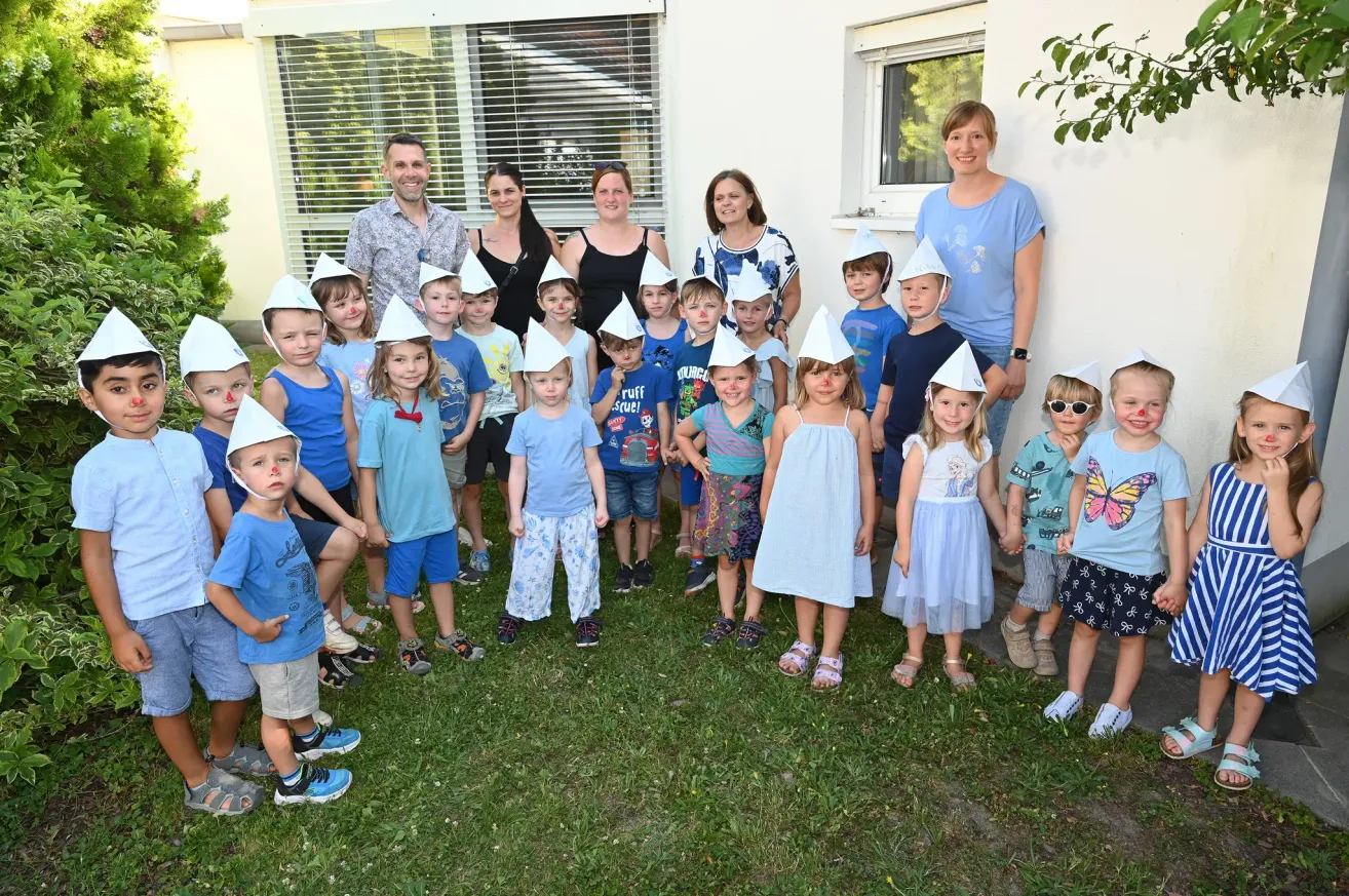 Eine Gruppe von Kindern und Erwachsenen in blauen Hemden und Hüten steht vor einem Gebäude. Einige haben rote Flecken auf den Wangen. Sie scheinen für ein Foto zu posieren.