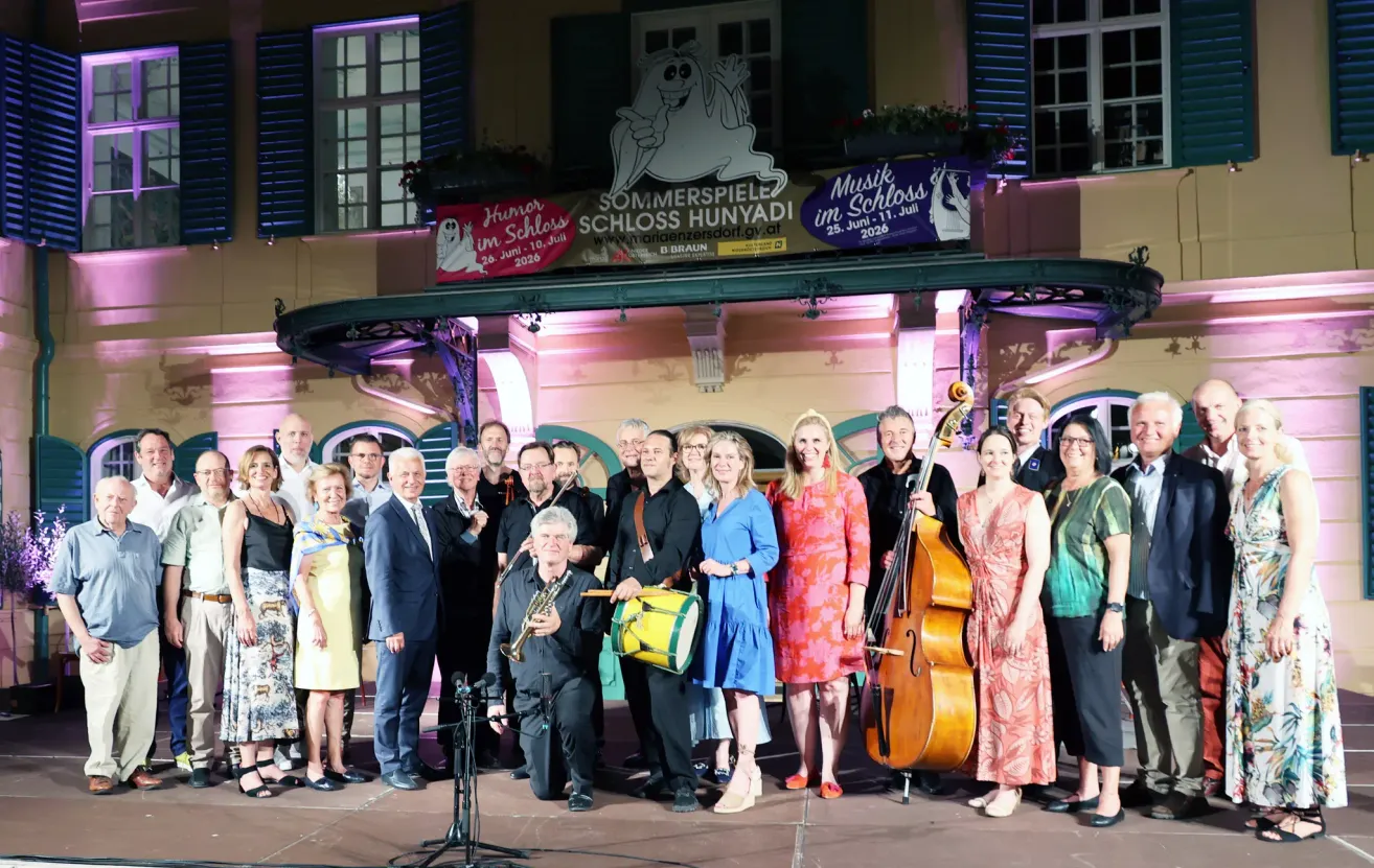 Eine Gruppe von Musikern und Offiziellen posiert für ein Foto vor einer dekorierten Bühne. Die Bühne zeigt Banner, die für Veranstaltungen im Juni 2026 werben.