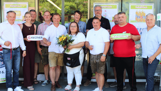 Eine Gruppe von Menschen steht vor einem Geschäft. Einige halten Schilder, darunter eines mit dem Namen Kastner. Sie tragen Freizeitkleidung, einige in traditioneller Tracht. Der Ort deutet auf eine Feier oder Werbeveranstaltung hin.