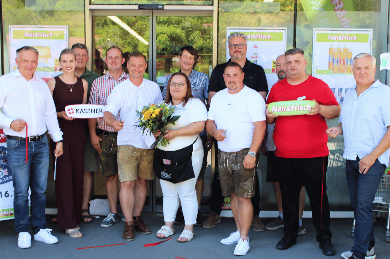 Eine Gruppe von Menschen steht vor einem Geschäft. Einige halten Schilder, darunter eines mit dem Namen Kastner. Sie tragen Freizeitkleidung, einige in traditioneller Tracht. Der Ort deutet auf eine Feier oder Werbeveranstaltung hin.