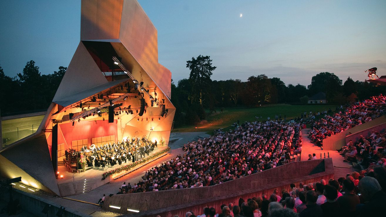 Ein Freiluft-Konzertsaal in der Dämmerung mit einem großen Publikum, das ein Symphonieorchester beobachtet. Die Bühne ist hell beleuchtet und das Publikum ist von roten Lichtern beleuchtet.