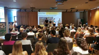 Ein Vortrag im Klassenzimmer mit einer Projektionsleinwand, die ein Projekt zur Zukunft des Klimas zeigt. Drei Personen stehen davor, und das Publikum sitzt in Reihen.