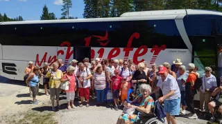 Eine Gruppe von Menschen, darunter eine Frau im Rollstuhl, steht vor einem großen Bus. Sie sind alle lässig gekleidet. Bäume sind im Hintergrund sichtbar.