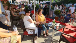 Zwei Frauen sitzen zusammen auf Stühlen im Freien, eine trägt eine Brille und ein weißes T-Shirt, die andere ein gelbes Oberteil. Ein Tisch mit einem Topf steht zwischen ihnen. Viele Menschen sind versammelt, einige stehen und andere sitzen. Bäume und Gebäude sind im Hintergrund.