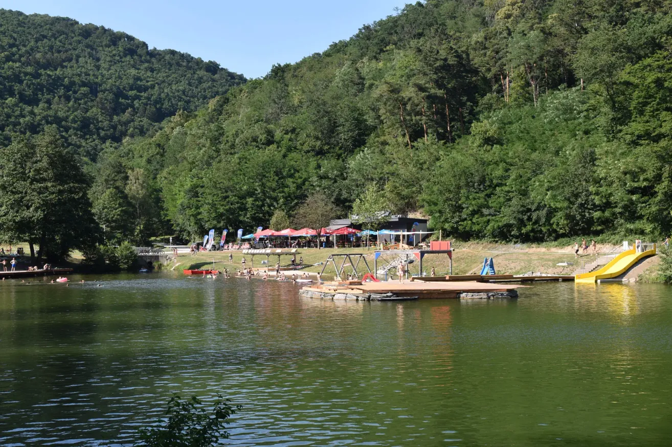 A lake with a wooden dock, boats, and people swimming. Green trees and mountains are in the background.