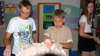 Zwei Jungen in einem Raum mit einer weißen Skulptur, einer mit einem Armband am Handgelenk. Ein farbenfrohes Bild ist an der Wand.