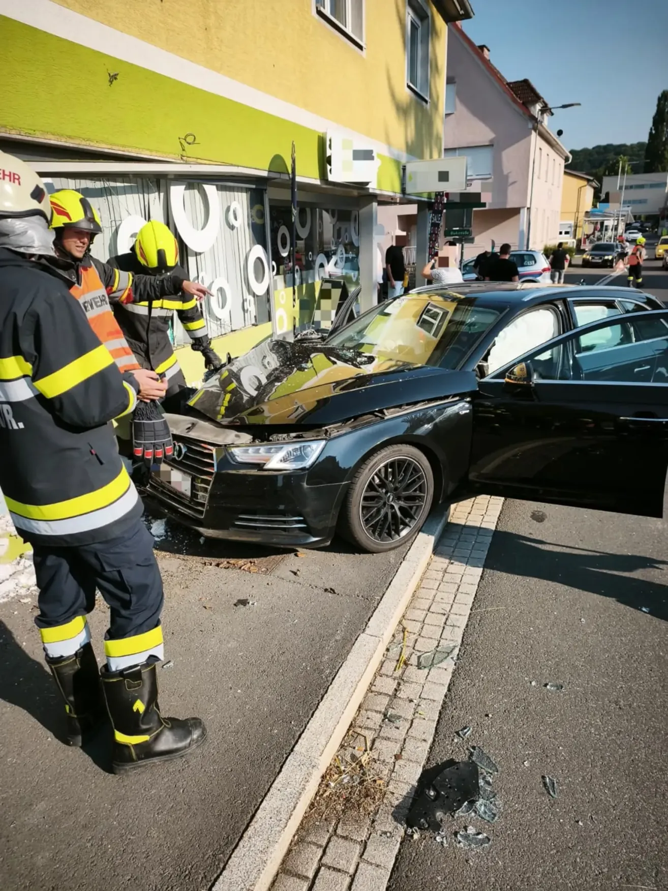 Feuerwehrleute inspizieren ein schwarzes Auto mit zerbrochener Front, das auf einer Straße geparkt ist, mit Leuten umher. Ein Geschäft mit Graffiti und einem beschädigten Schaufenster ist hinter dem Auto.