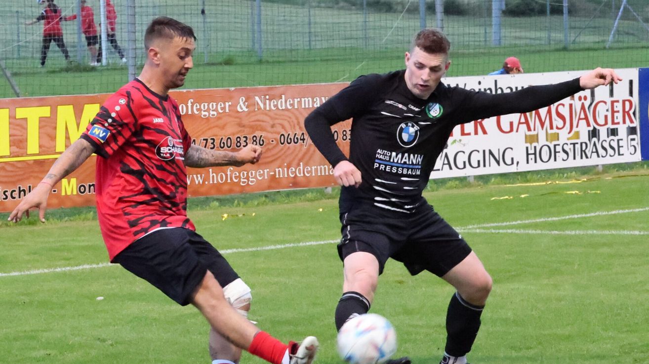 Zwei Fußballspieler in Aktion auf einem Feld. Einer in einem rot-schwarzen Trikot, der andere in Schwarz mit BMW-Logo. Ein Fußball liegt zwischen ihnen.