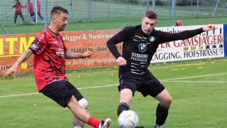 Zwei Fußballspieler in Aktion auf einem Feld. Einer in einem rot-schwarzen Trikot, der andere in Schwarz mit BMW-Logo. Ein Fußball liegt zwischen ihnen.