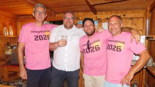 Vier Männer tragen rosafarbene T-Shirts mit der Nummer 2026. Zwei von ihnen lächeln, während einer einen Becher hält. Hinter ihnen ist eine Holzwand.