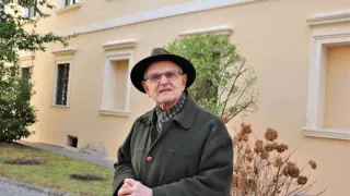 Ein älterer Mann mit Brille und Hut steht vor einem Gebäude mit einem Baum und Gras.