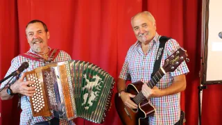 Zwei Männer lächeln vor einem roten Vorhang. Ein Mann spielt Akkordeon, der andere Gitarre.