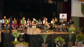 Eine Gruppe von Musikern spielt auf einer Bühne mit Instrumenten wie Cello, Tuba und Gitarre. Einige sitzen auf Stühlen, andere stehen. Eine Fahne ist an der Rückwand.