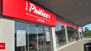 Die Storefront von Philipp in Hartberg, mit einem roten Schild mit weißen Buchstaben und Glastüren, die umliegende Autos und Grünflächen reflektieren.