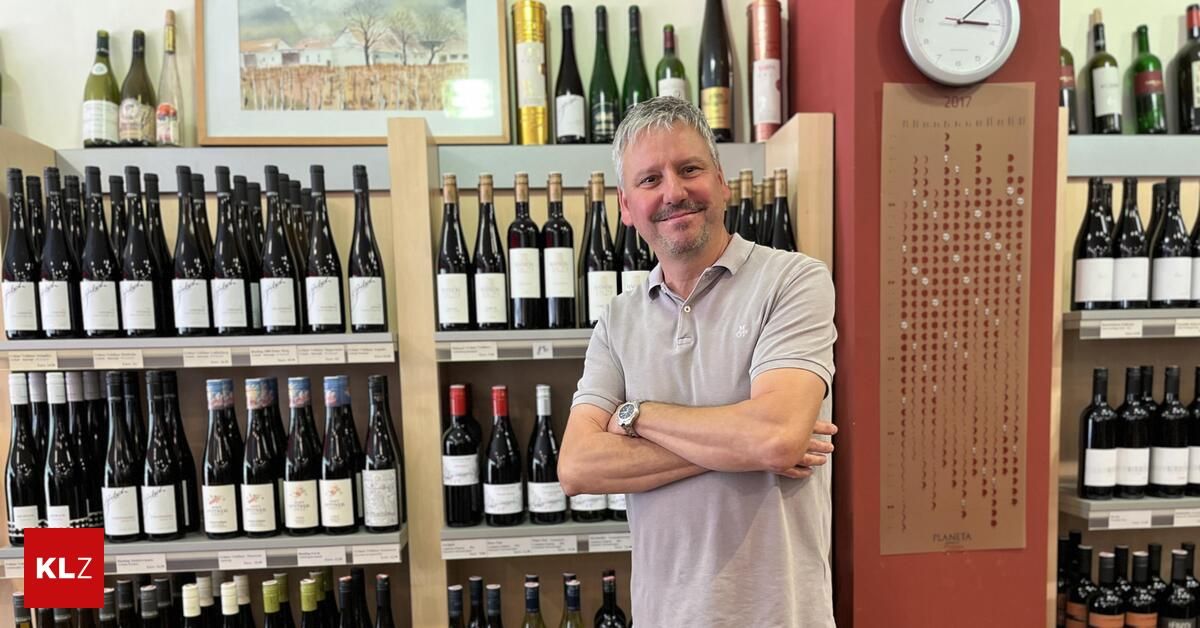 Ein Mann steht in einem Weinladen und lächelt mit verschränkten Armen. Hinter ihm stehen Regale voller Weinflaschen. Über den Regalen befindet sich ein Bild und eine Uhr an der Wand.