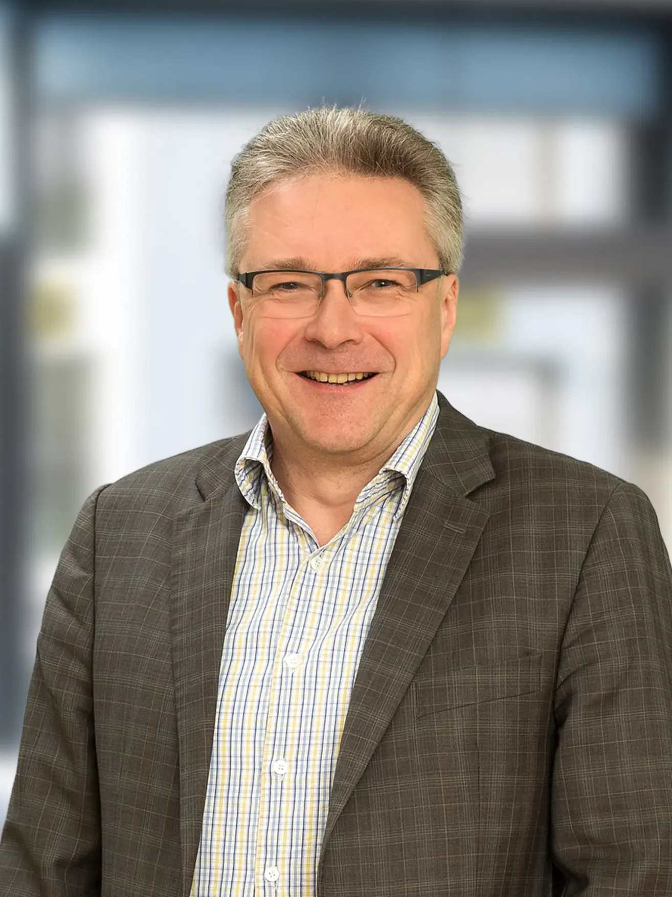 Ein lächelnder Mann in einer Anzugjacke und mit Brille, mit einem karierten Hemd, steht vor einem verschwommenen Hintergrund.