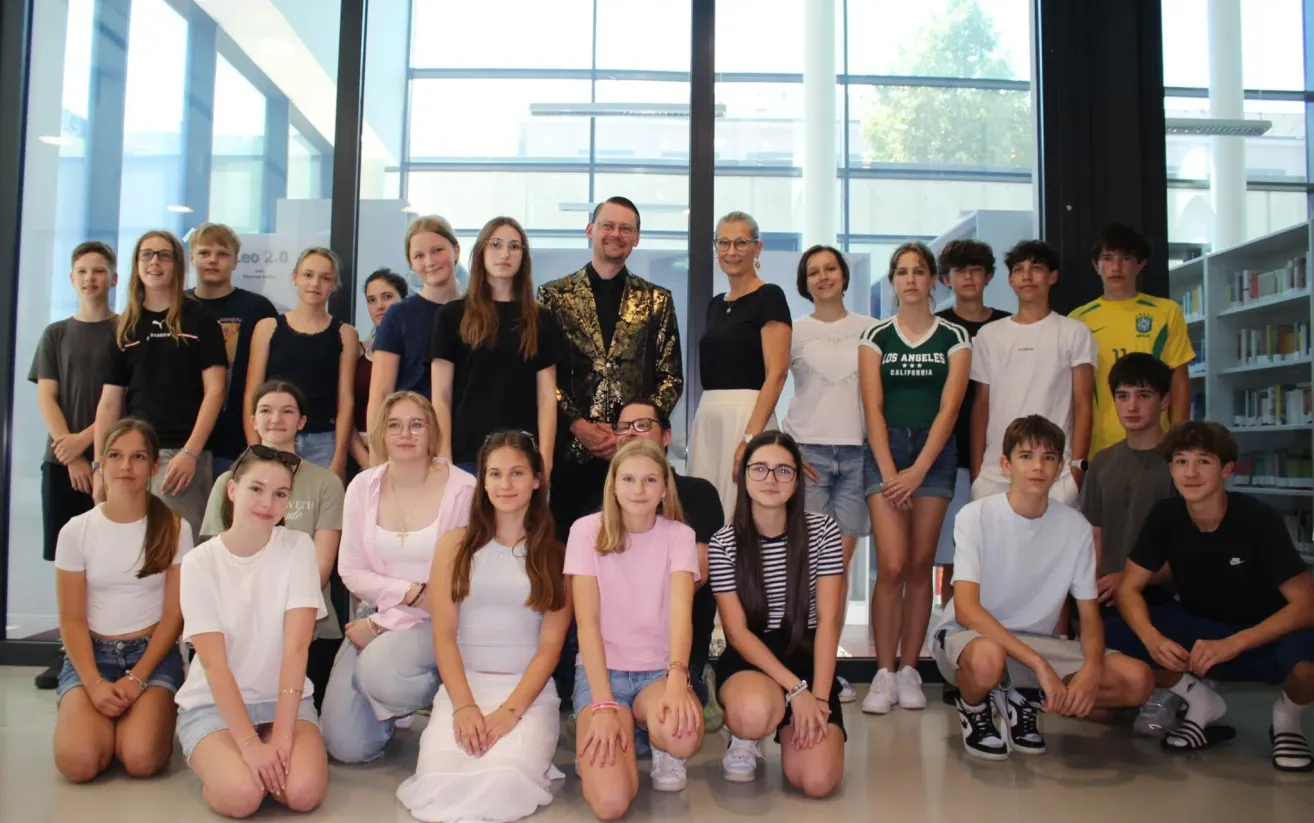 Eine Gruppe von Menschen in einem Studio, einige sitzen und einige stehen, in lässiger Kleidung, posiert für ein Foto. Der Hintergrund ist eine Glaswand mit Reflexionen.