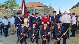Eine Gruppe von Männern in Uniformen posiert für ein Foto vor einer Steinmauer, einige halten Stöcke mit Blumen. Dahinter sind andere Leute.