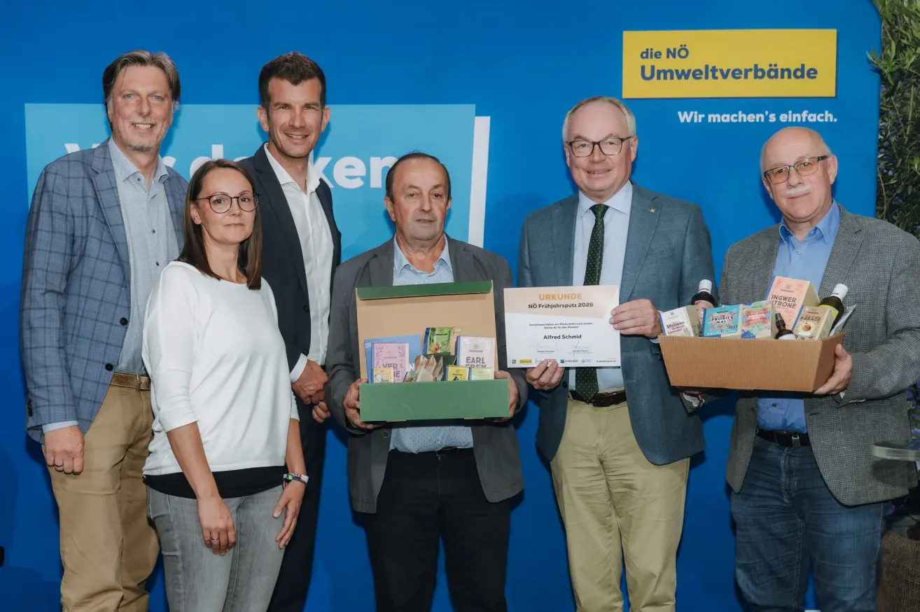 Five people stand in front of a blue backdrop. They hold a box and a certificate. The man in the middle smiles. The man on the right holds a box with products. The man on the left holds a box with books.