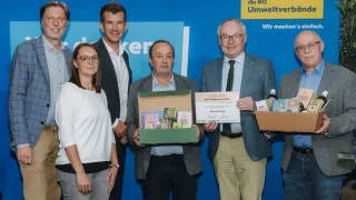 Fünf Personen stehen vor einem blauen Hintergrund. Sie halten eine Box und ein Zertifikat. Der Mann in der Mitte lächelt. Der Mann auf der rechten Seite hält eine Box mit Produkten. Der Mann auf der linken Seite hält eine Box mit Büchern.