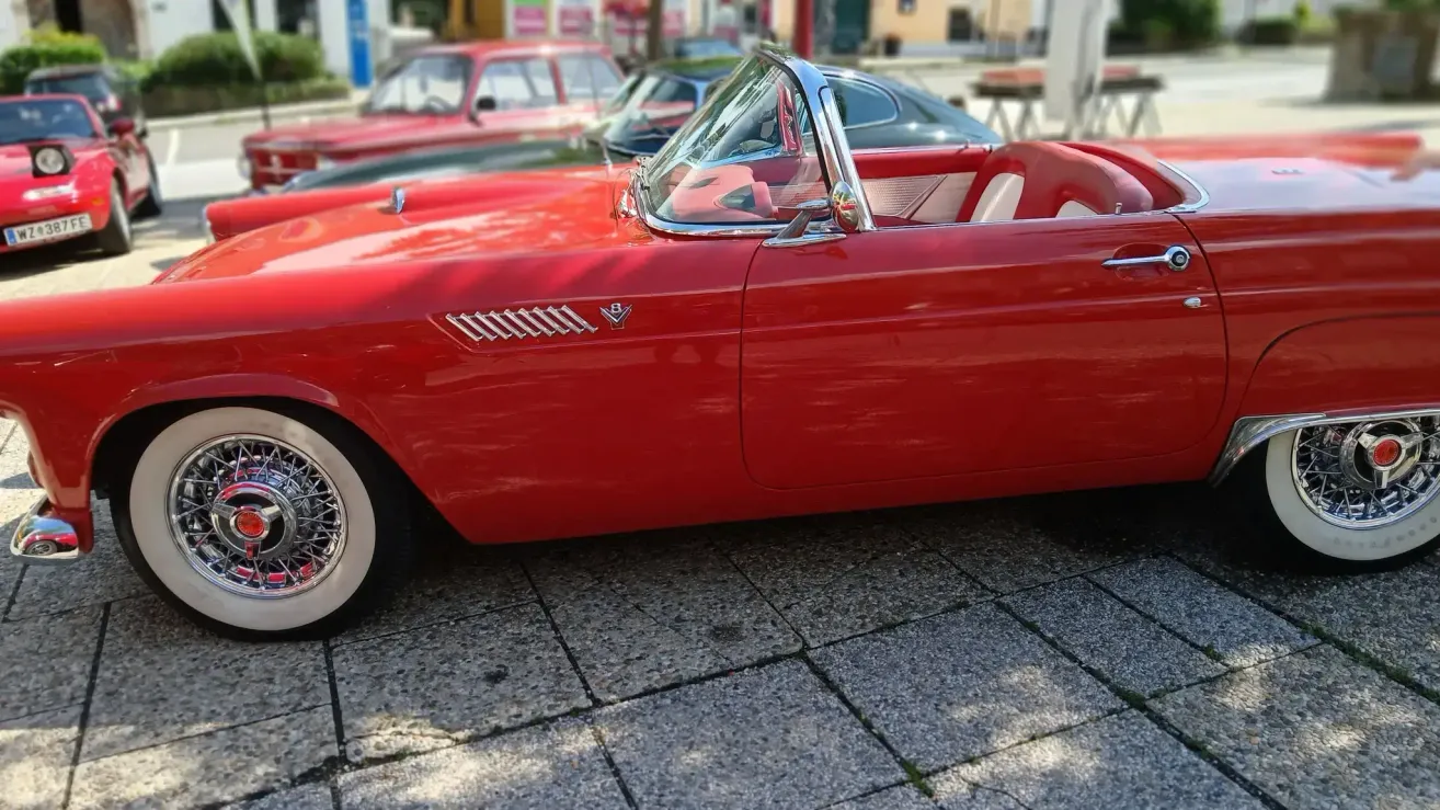 Ein roter Vintage Ford Thunderbird Cabriolet auf einem gepflasterten Bereich geparkt, mit einem roten Auto und einem schwarzen Auto dahinter.