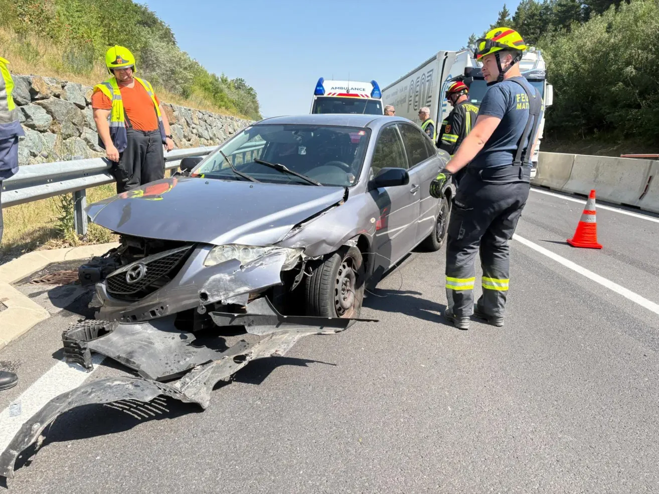 Notfallhelfer stehen neben einem beschädigten Auto. Einer in reflektierender Kleidung inspiziert die Front. In der Nähe geht eine Person in einem Helm am Straßenrand entlang. Dahinter sind Notfallfahrzeuge und ein Geländer sichtbar.