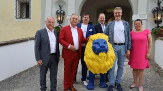 Fünf Männer stehen draußen vor einem Gebäude, einer hält eine gelbe und blaue Löwenstatue. Sie sind in Anzug gekleidet.