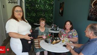 Vier Frauen sitzen um einen Tisch mit Weinglas und Desserts. Die Frau links hält eine schwarze Tasche. Eine Frau in der Mitte trägt eine Halskette. Eine Frau rechts trägt eine Uhr.