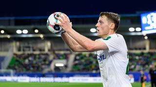 Ein Fußballspieler fängt einen Fußball in der Luft auf einem Stadionfeld bei Nacht.