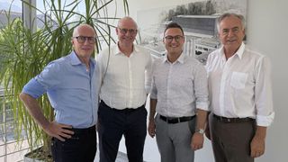 Vier Männer stehen zusammen vor einer Wandmalerei. Sie tragen alle formelle Kleidung. Der Mann auf der linken Seite trägt eine Brille und ein blaues Hemd. Der Mann neben ihm trägt ein weißes Hemd. Der Mann rechts trägt ein weißes Hemd und eine Uhr.