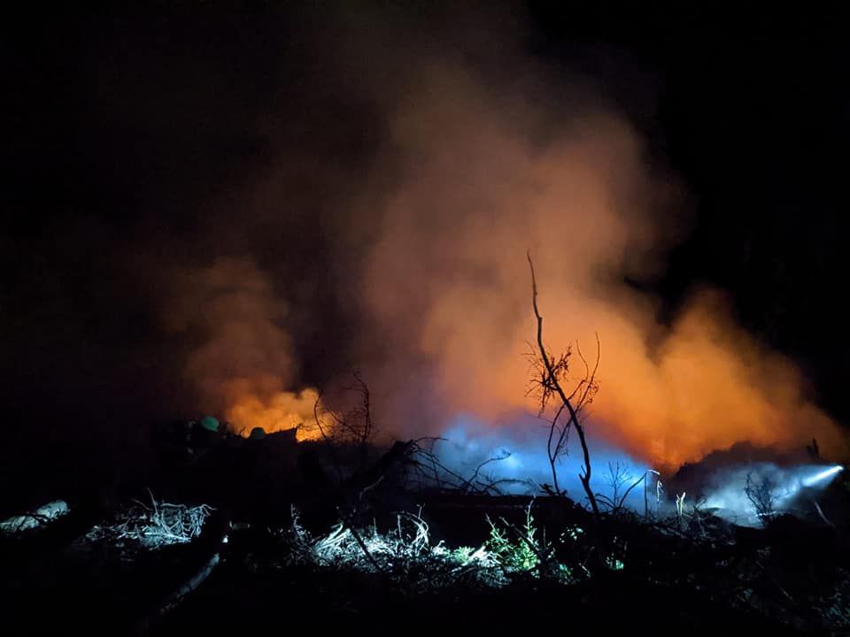 A nighttime landscape shows a fire with thick smoke and blue and orange flames, illuminating the dark surroundings.