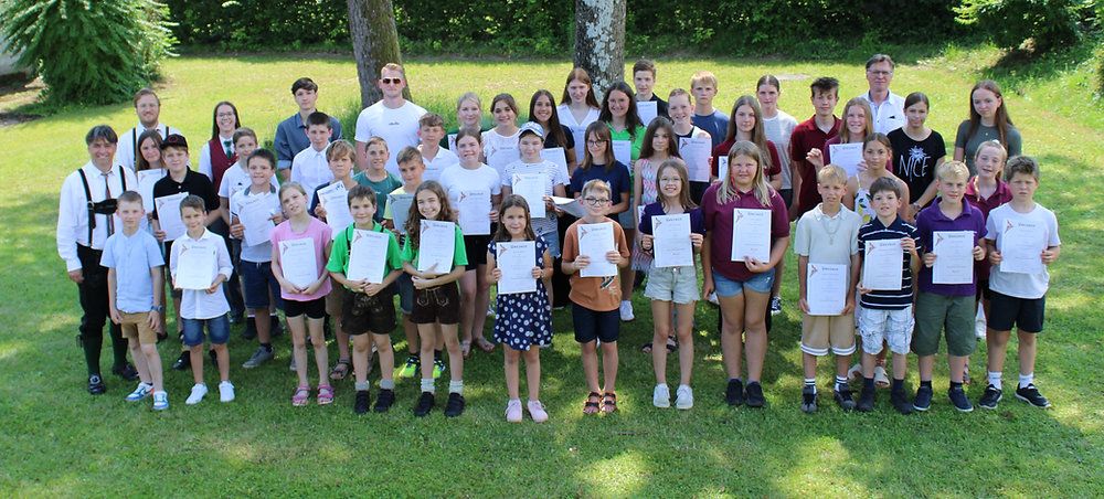 Eine große Gruppe von Kindern und Erwachsenen posiert für ein Foto auf einem Rasen, während sie Urkunden halten. Sie lächeln und tragen Freizeitkleidung.