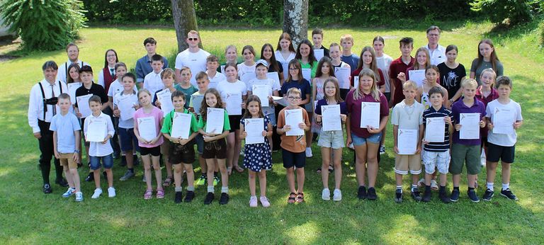 Eine große Gruppe von Kindern und Erwachsenen posiert für ein Foto auf einem Rasen, während sie Urkunden halten. Sie lächeln und tragen Freizeitkleidung.
