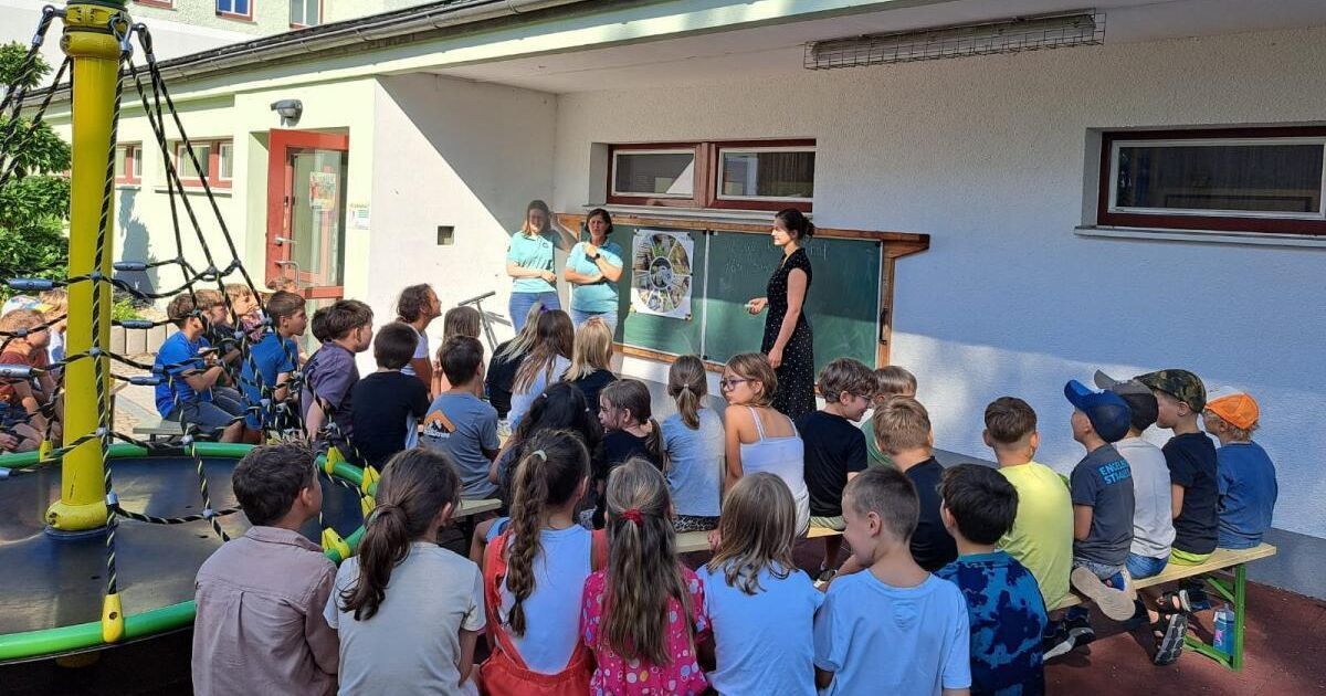 Zwei Frauen stehen vor einem Klassenzimmer mit einer Tafel und einem Plakat. Viele Kinder sind um sie herum versammelt. Das Klassenzimmer befindet sich im ersten Stock eines Gebäudes.