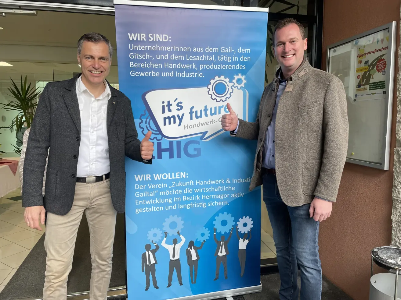 Two men stand next to a blue banner with a slogan and illustrations of people raising their hands. The banner says 'it's my future'. One man gives a thumbs up.