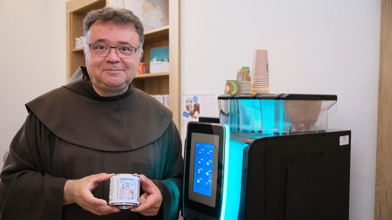 Ein Mann in einer braunen Robe hält eine Dose mit einem Bild einer Kirche. Er steht neben einer Maschine mit blauen Lichtern.
