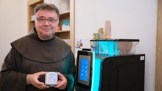 Ein Mann in einer braunen Robe hält eine Dose mit einem Bild einer Kirche. Er steht neben einer Maschine mit blauen Lichtern.