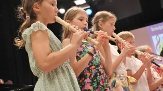 Vier junge Mädchen spielen Flöten in einem Konzertsaal. Das Mädchen vorne trägt ein Blumenkleid, während die anderen auch Blumenkleider tragen.