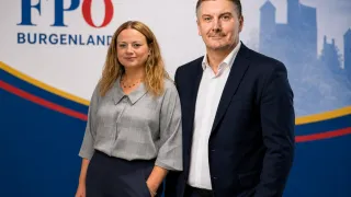 Ein Mann und eine Frau stehen Seite an Seite vor einem PO-Schild. Die Frau trägt eine graue Bluse, der Mann einen dunklen Anzug.