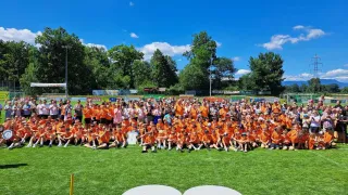 Eine große Gruppe von Menschen in orangefarbenen Shirts posiert für ein Foto auf einem Rasenfeld. Hinter ihnen stehen Bäume, eine Anzeigetafel und ein Banner.